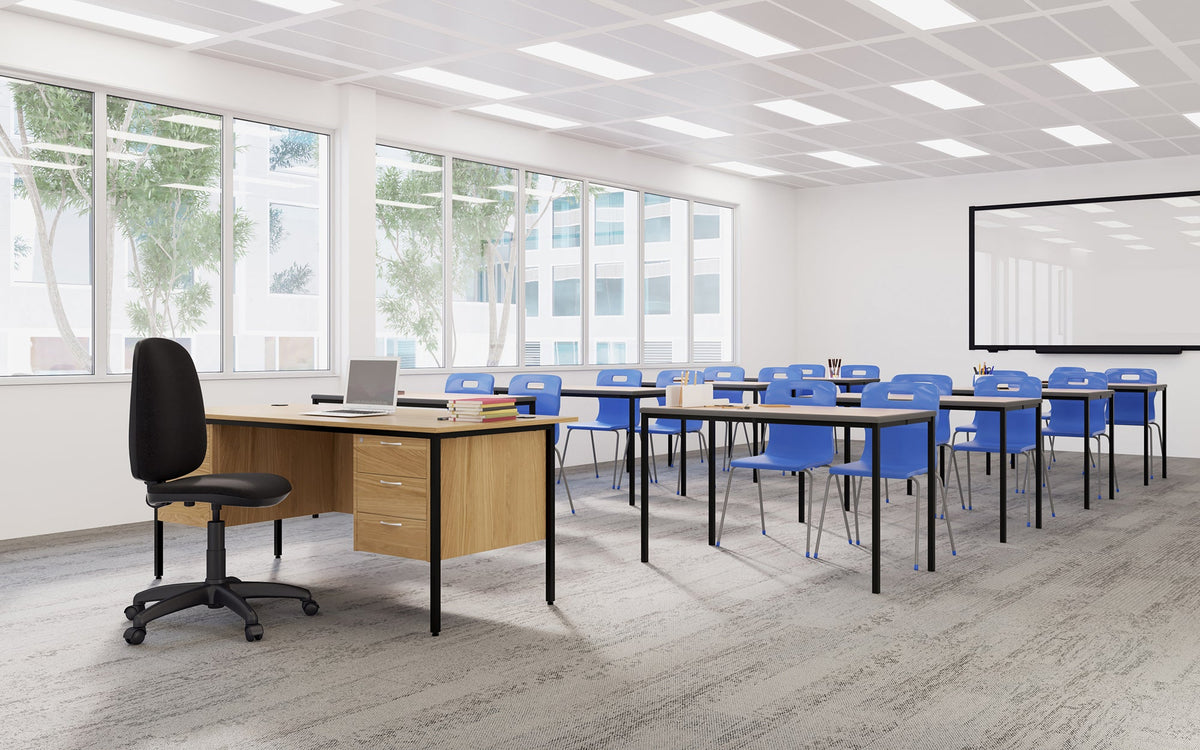 A modern classroom or meeting space with rows of rectangular desks and blue chairs, plus a wooden office desk with a black swivel chair near large windows.