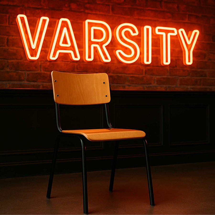 Neon 'Varsity' sign above a wooden chair against a brick wall.